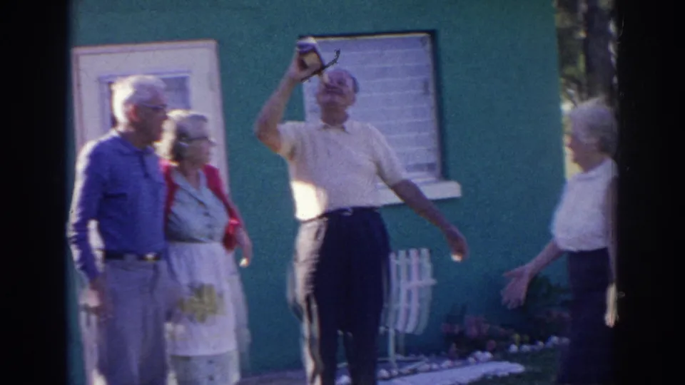 Near the end of the 1966 Norfolk clip, the man holds the bottle overhead while older adults gather around him outdoors.