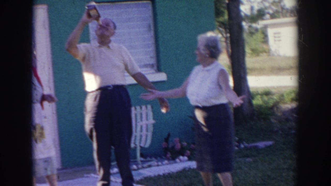 Older man drinks from a bottle while a woman gestures beside him outdoors.