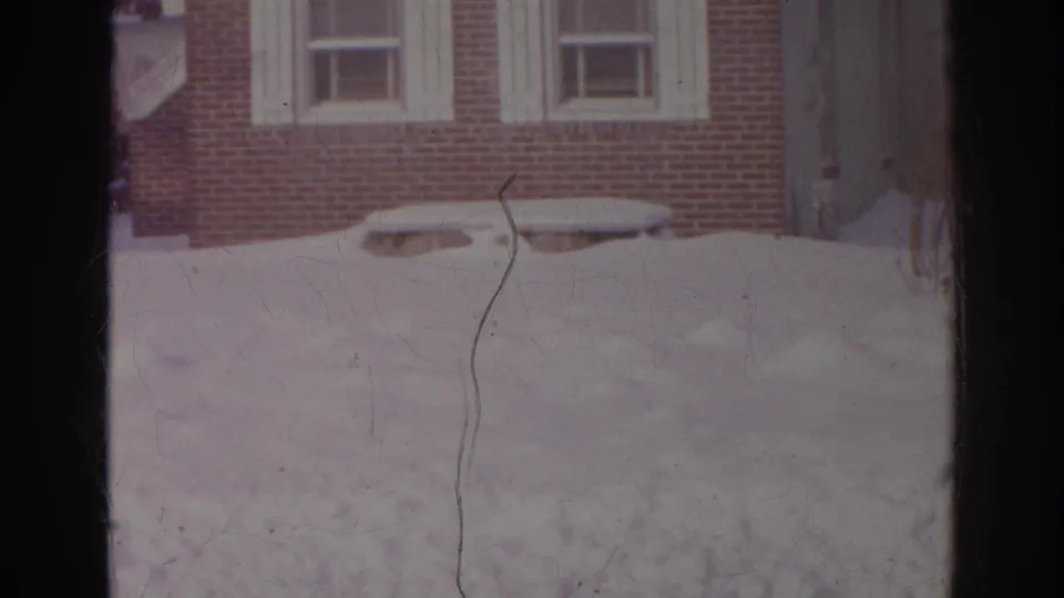 The final frame holds on snow outside a brick residential facade, with scratches and frame bars visible in the 8mm scan.