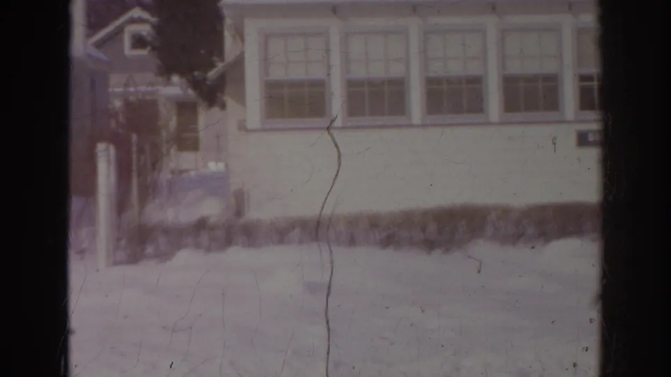 1966 Norfolk winter frame showing a snow-covered residential yard, pale house windows, shrubs, and a partly visible parked car.