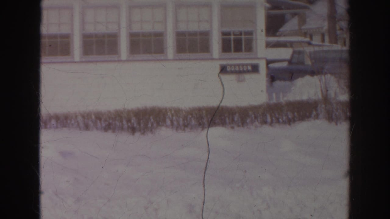 1966 Norfolk winter exterior with a DOBSON sign on a white building, snowbanks, hedge, and a dark parked car at the edge of frame.