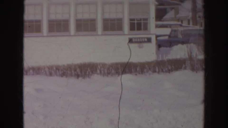 1966 Norfolk winter exterior with a DOBSON sign on a white building, snowbanks, hedge, and a dark parked car at the edge of frame.