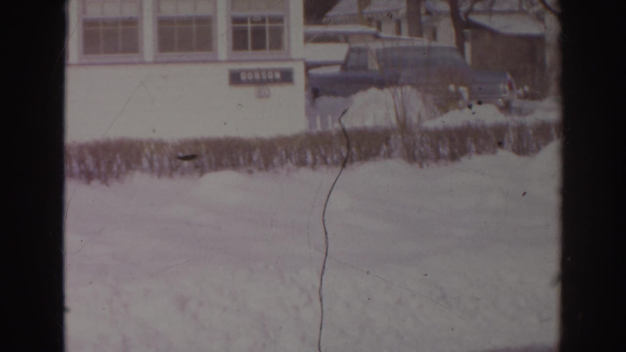 1966 Norfolk 8mm still showing a dark parked car beside a snow-covered hedge and the white DOBSON building.