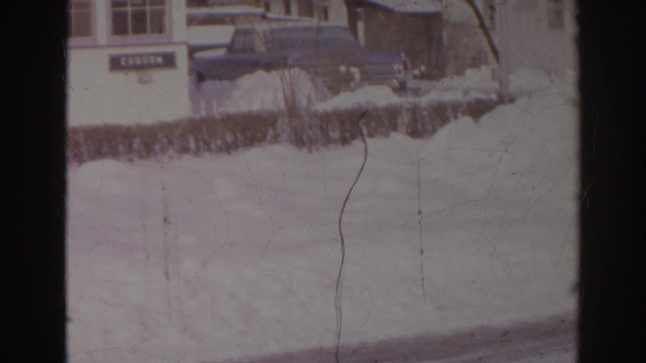 1966 Norfolk winter frame with a dark car parked beside the DOBSON-marked building and snow covering the foreground.