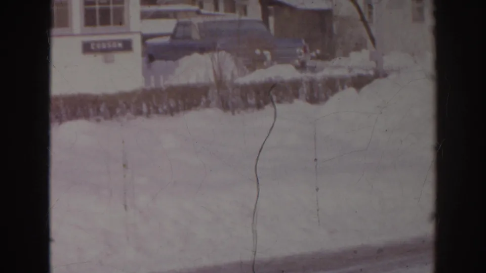 1966 Norfolk winter frame with a dark car parked beside the DOBSON-marked building and snow covering the foreground.