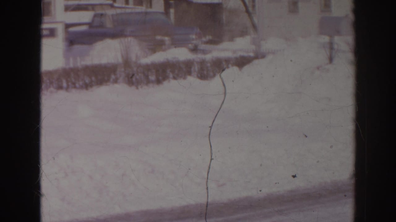Snow-covered residential street with parked cars and houses in the background.