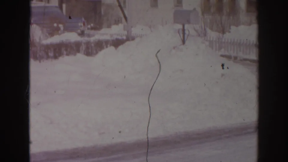 1966 Norfolk suburban winter view with a snowbank, roadside mailbox, fence, and houses beyond the street.