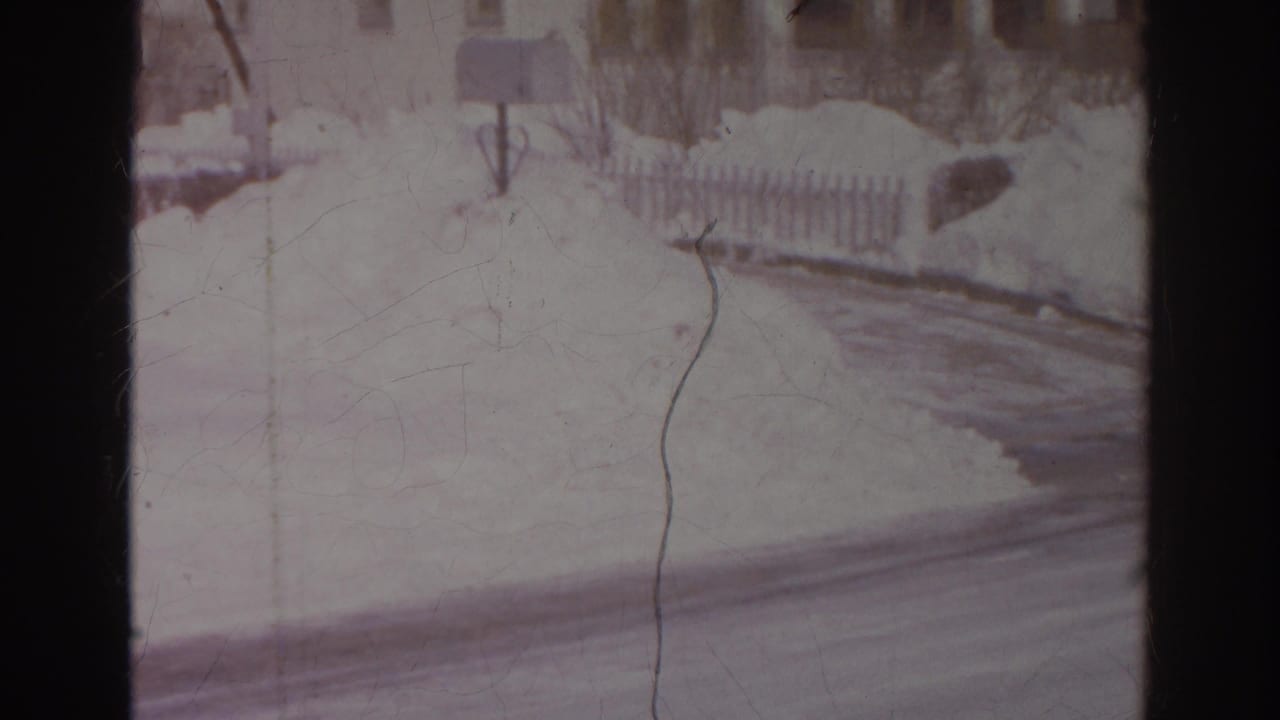 Home-movie view of a snowy Norfolk residential road curve with a mailbox, fence, snowbanks, and nearby houses.