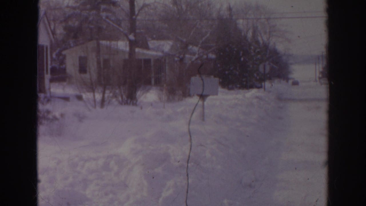 1966 Norfolk winter frame showing a snow-covered yard, mailbox, detached house, bare trees, and road edge.