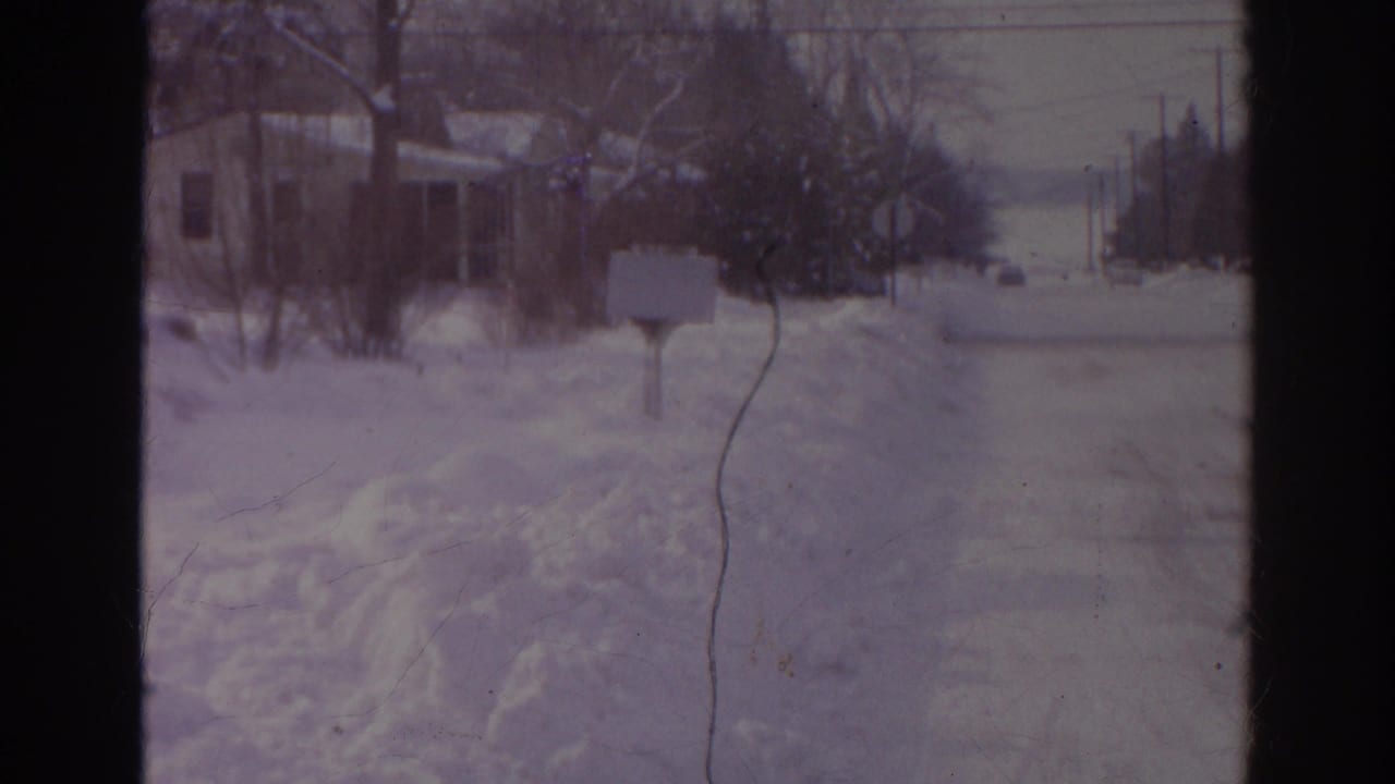 Snow-covered residential street with snowbanks, bare trees, a mailbox, and houses in the distance.