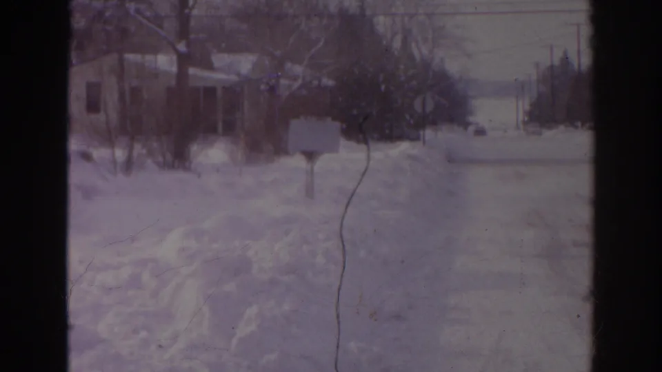 1966 Norfolk home-movie frame showing a snowy residential road bordered by snowbanks, bare trees, a mailbox, and distant houses.