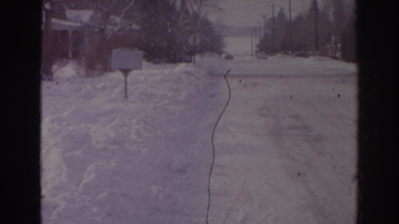1966 Norfolk frame looking down a snow-covered street toward an intersection with roadside signs, utility poles, and heavy snowbanks.