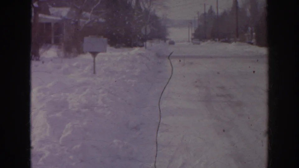 1966 Norfolk frame looking down a snow-covered street toward an intersection with roadside signs, utility poles, and heavy snowbanks.
