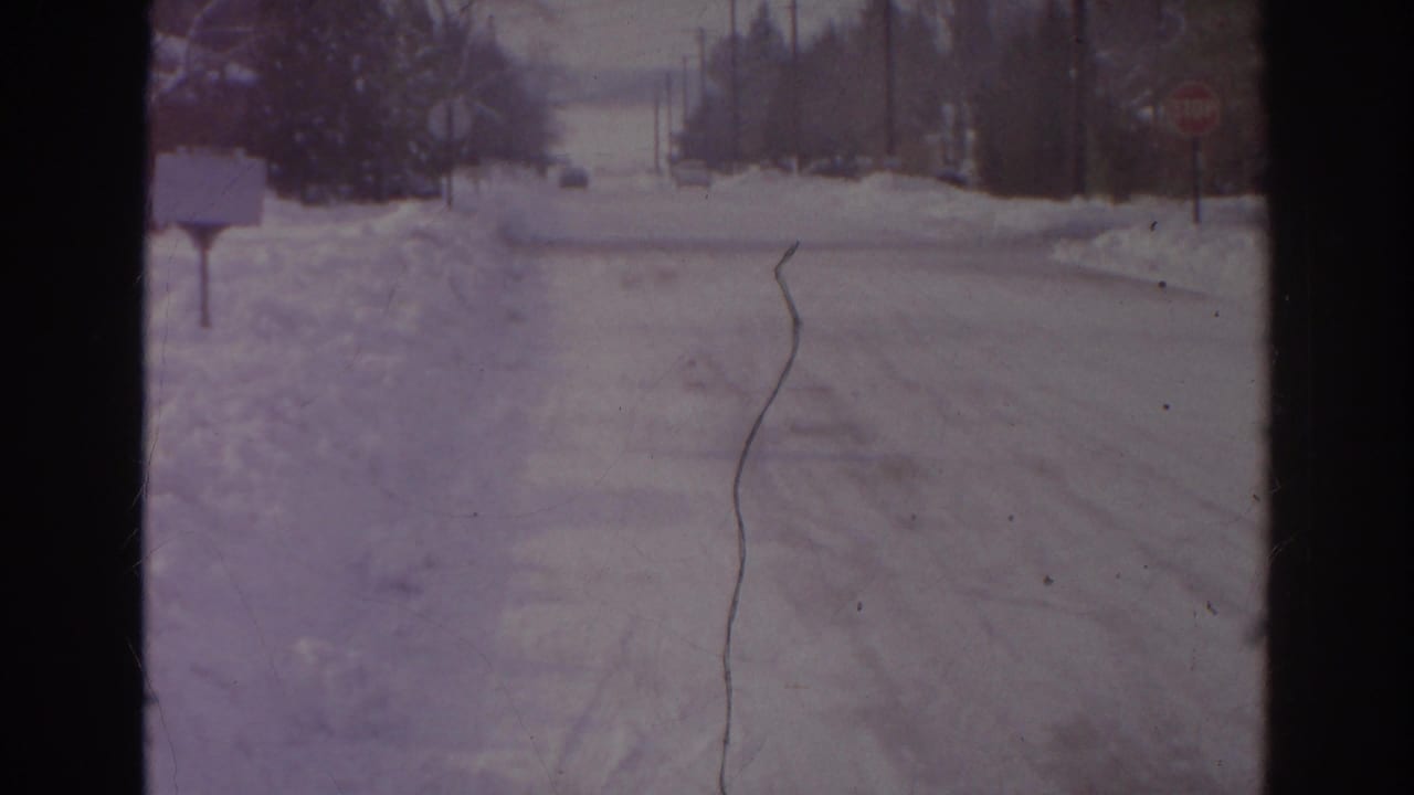 1966 Norfolk home-movie view of a snow-covered street with a mailbox at left, a distant stop sign, snowbanks, and roadside utility poles.