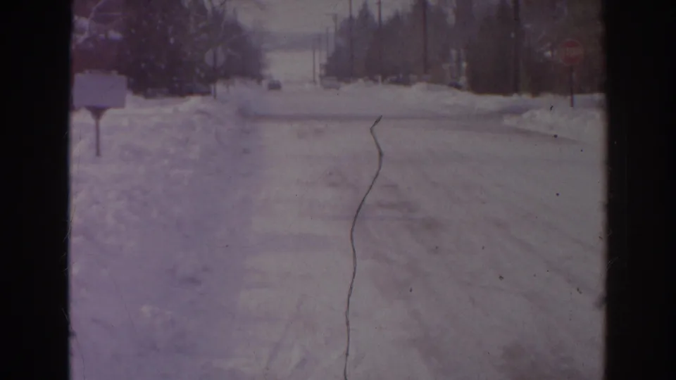 1966 Norfolk home-movie view of a snow-covered street with a mailbox at left, a distant stop sign, snowbanks, and roadside utility poles.