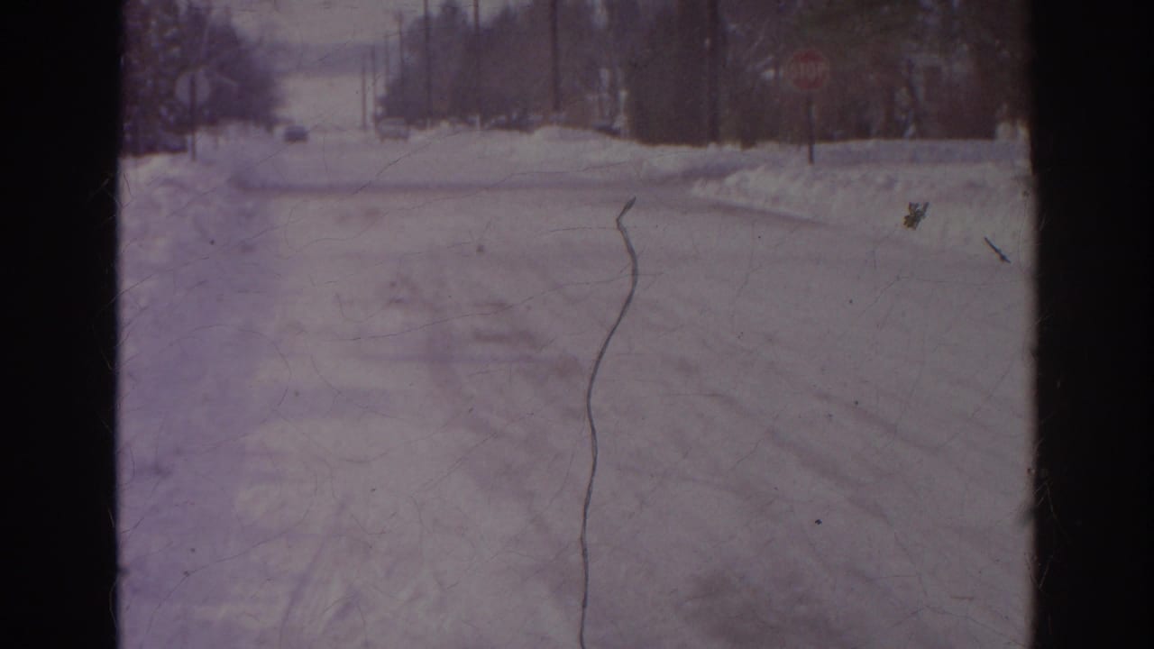 1966 Norfolk frame showing a snowy residential road with visible tire tracks, snowbanks, stop signs, bare trees, and utility poles.
