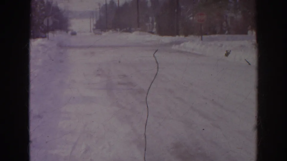 1966 Norfolk frame showing a snowy residential road with visible tire tracks, snowbanks, stop signs, bare trees, and utility poles.