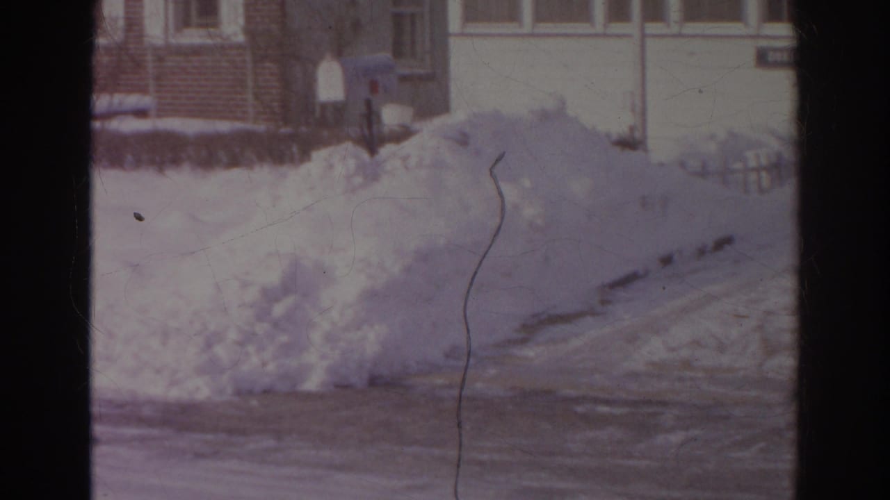 Norfolk home-movie still from 1966 showing a roadside snow pile, house fronts, mailboxes, and a dark vertical film divider.