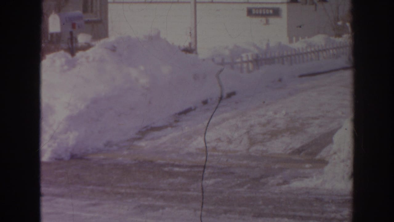 1966 Norfolk frame with a snowbank, icy roadway, stop sign, mailboxes, and residential house exteriors in soft 8mm color.