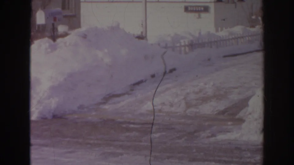1966 Norfolk frame with a snowbank, icy roadway, stop sign, mailboxes, and residential house exteriors in soft 8mm color.