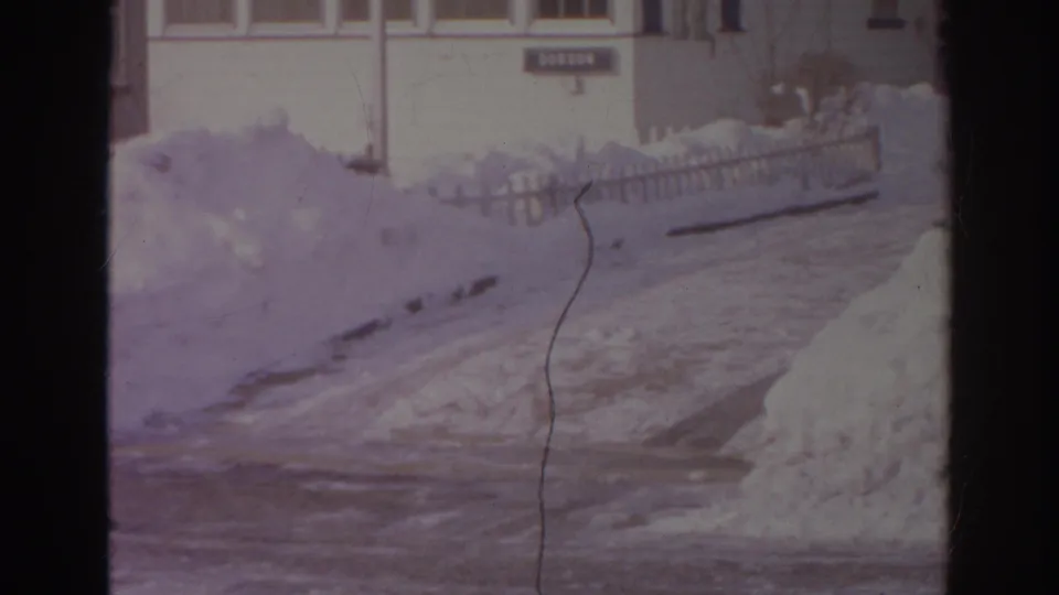1966 Norfolk home-movie frame showing deep snow along a residential street, with picket fencing and house fronts beyond it.
