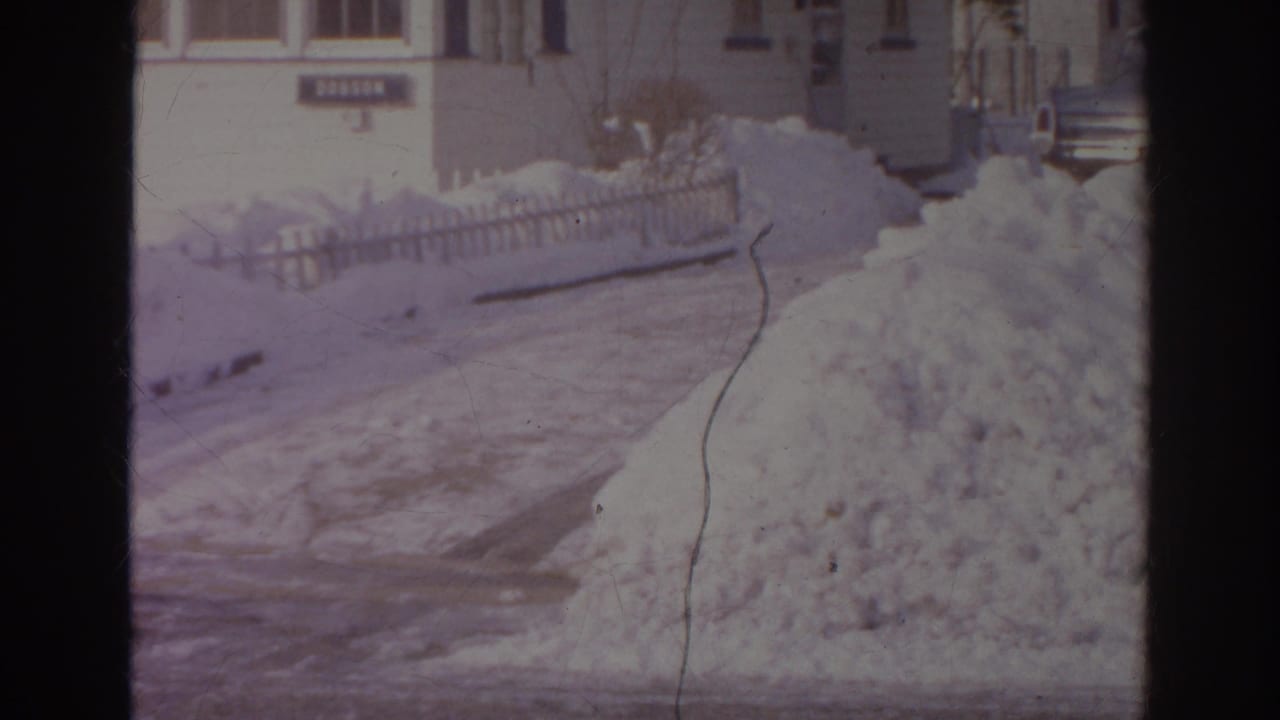 1966 Norfolk frame with piled snow beside the road, low fencing, bare shrubs, and residential houses in the background.