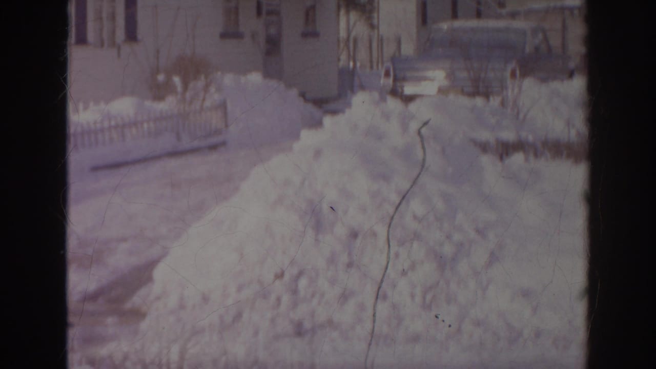 1966 Norfolk home-movie frame showing a high snow pile, a partly visible parked car, fencing, and nearby houses.
