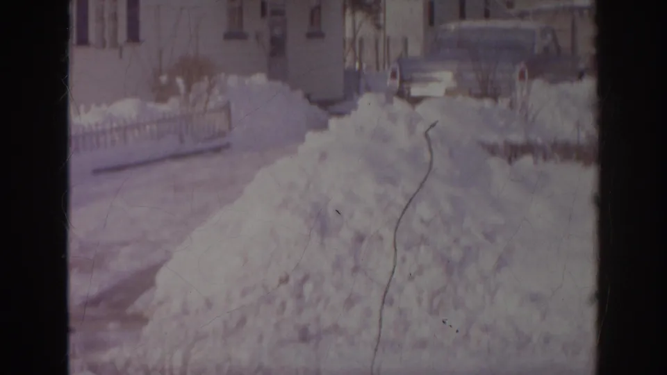 1966 Norfolk home-movie frame showing a high snow pile, a partly visible parked car, fencing, and nearby houses.
