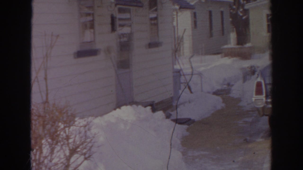 1966 Norfolk frame looking toward a house exterior and walkway bordered by deep snow and winter shrubs.