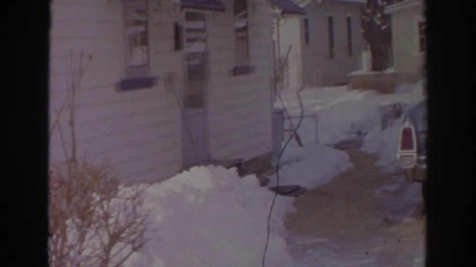 1966 Norfolk frame looking toward a house exterior and walkway bordered by deep snow and winter shrubs.