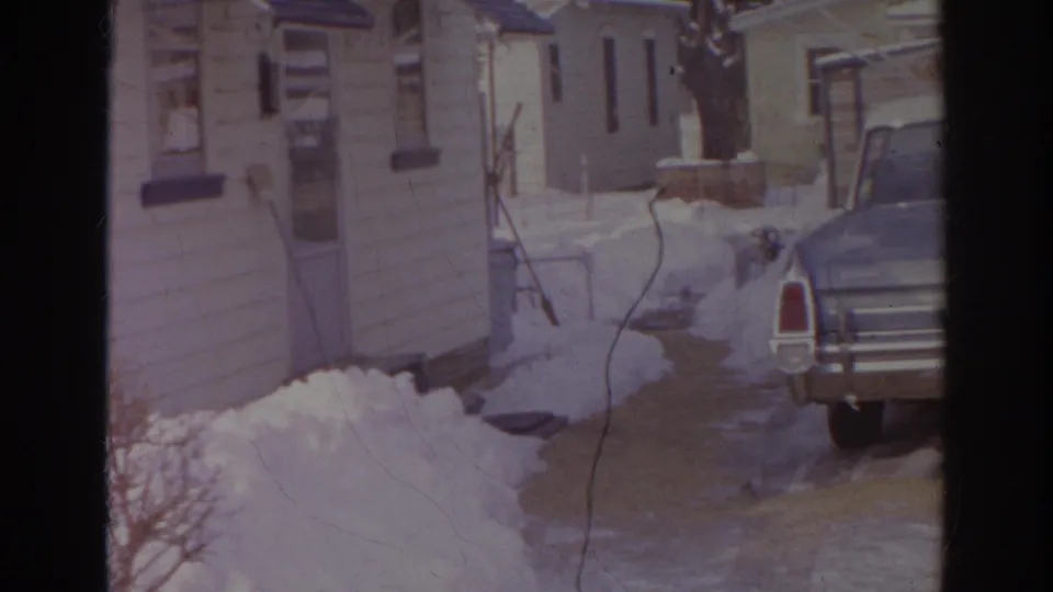 1966 Norfolk home-movie frame of a blue American sedan in a snowy driveway beside tan residential buildings.