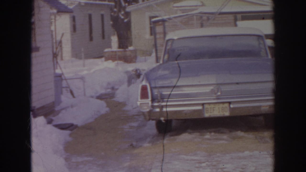 Rear view of a blue sedan parked in a snow-covered Norfolk driveway, with tan houses and bare trees behind it.