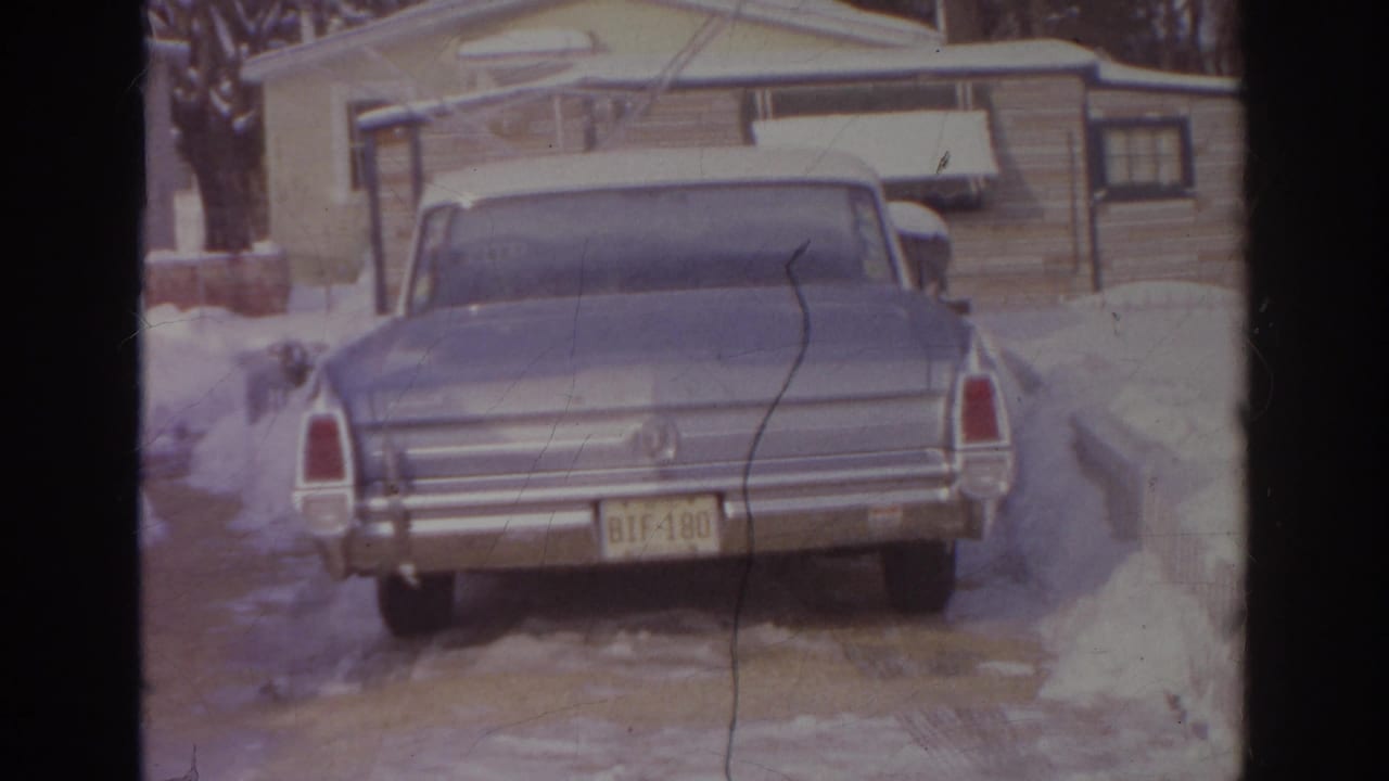 Closer 1966 8mm view of the blue car's rear, with a yellow plate, chrome bumper, snowbanks, and residential siding visible.