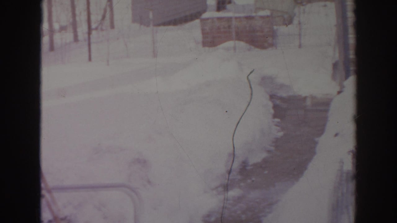 1966 Norfolk home-movie frame showing a shoveled path through snow toward a residential exterior and yard structures.
