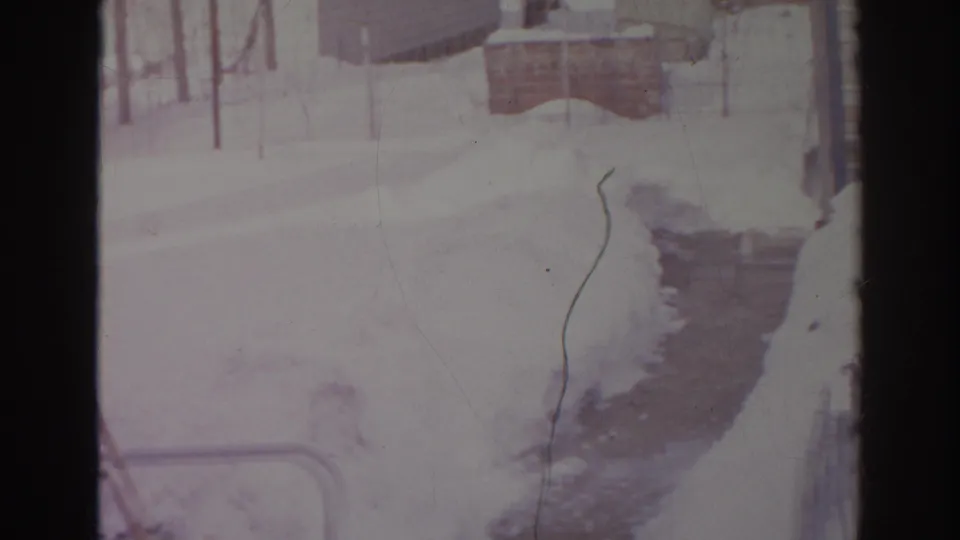 1966 Norfolk home-movie frame showing a shoveled path through snow toward a residential exterior and yard structures.