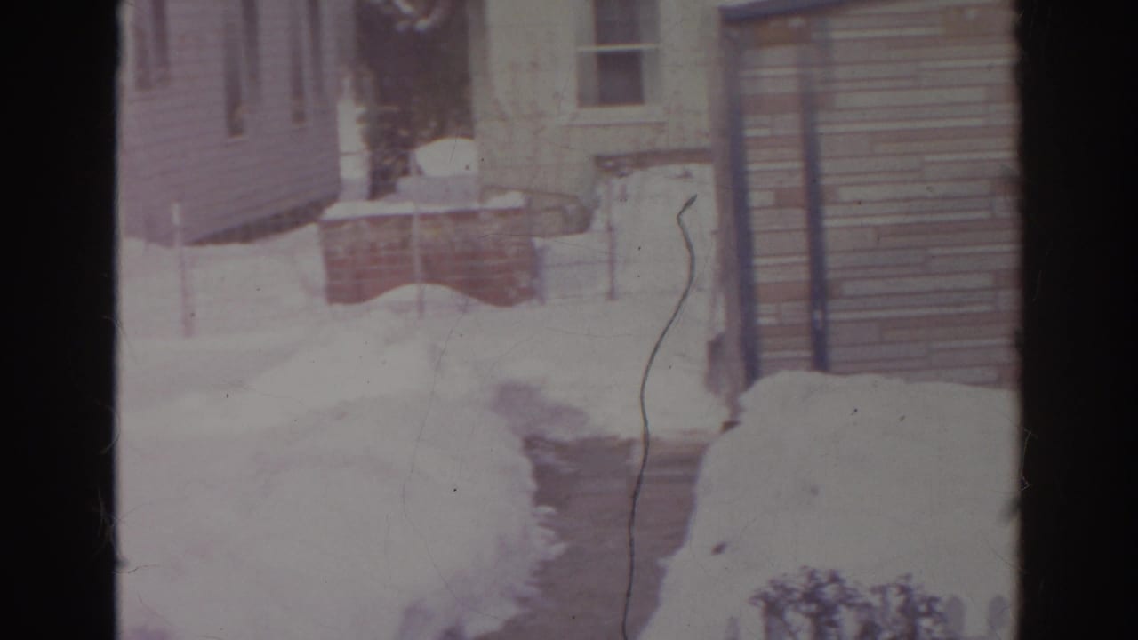 1966 Norfolk home-movie frame of a snow-lined walkway beside a residential exterior with siding, steps, and yard snowbanks.