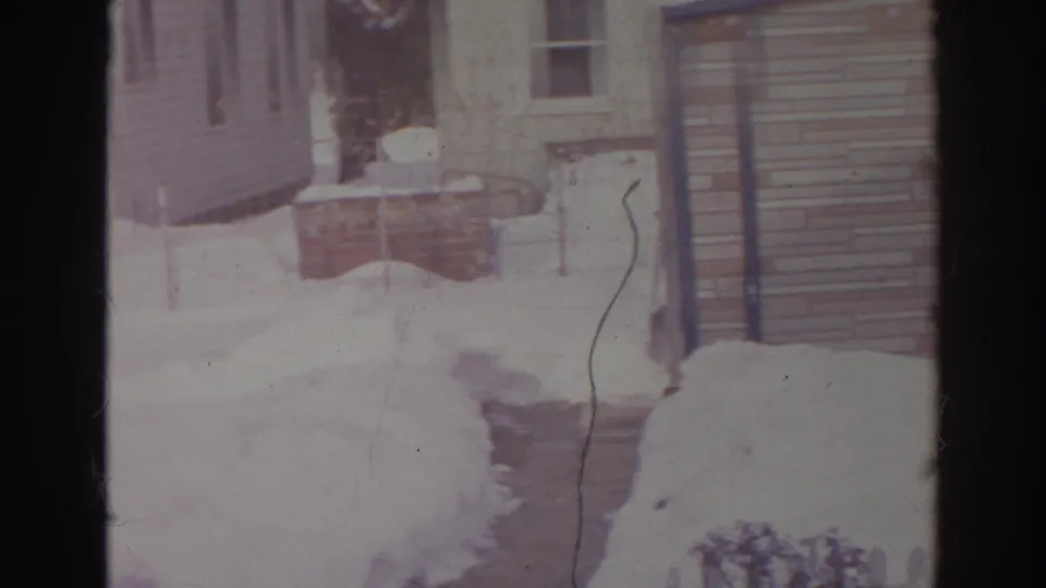 1966 Norfolk home-movie frame of a snow-lined walkway beside a residential exterior with siding, steps, and yard snowbanks.