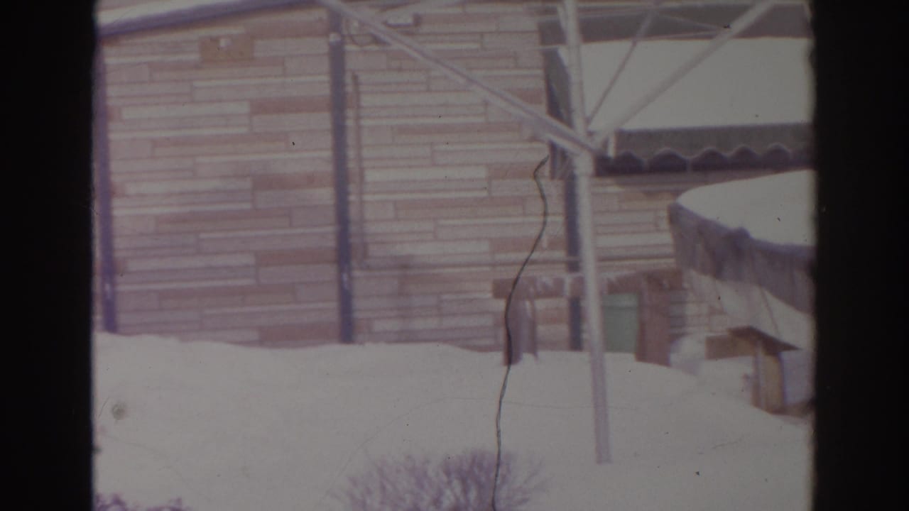 Snowy residential backyard in Norfolk, 1966, showing siding, patio posts, an awning edge, and shrubs under deep snow.