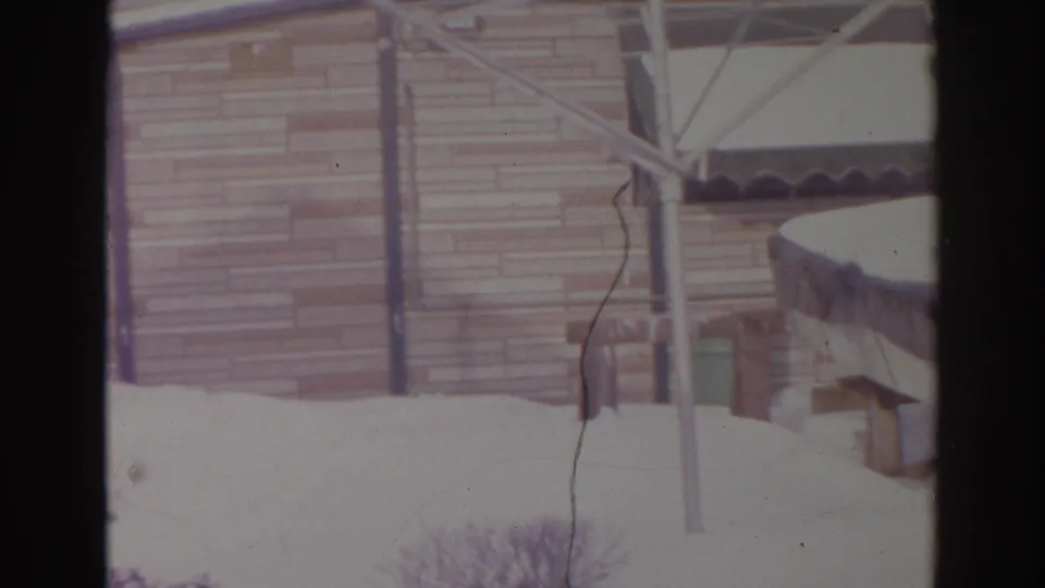 Snowy residential backyard in Norfolk, 1966, showing siding, patio posts, an awning edge, and shrubs under deep snow.