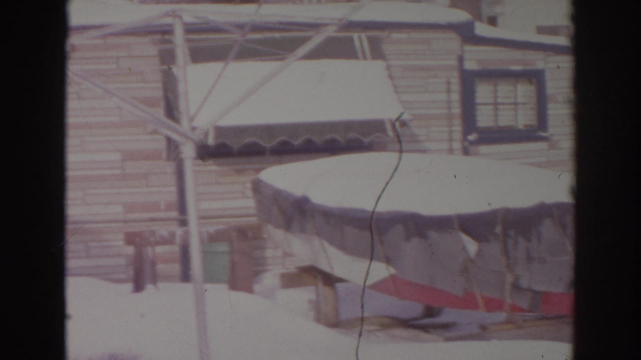 Snow-covered boat on a trailer beside a wooden-sided house.