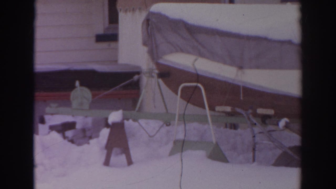 Snow-covered boat on a trailer beside a house, with a small wooden yard structure nearby.