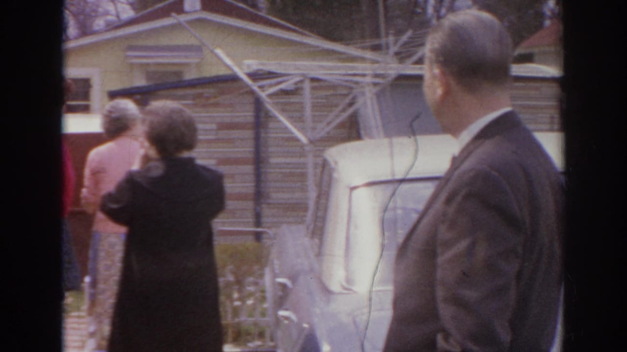 1966 Norfolk frame with a suited man in the foreground and adults conversing near a parked car in a residential yard.