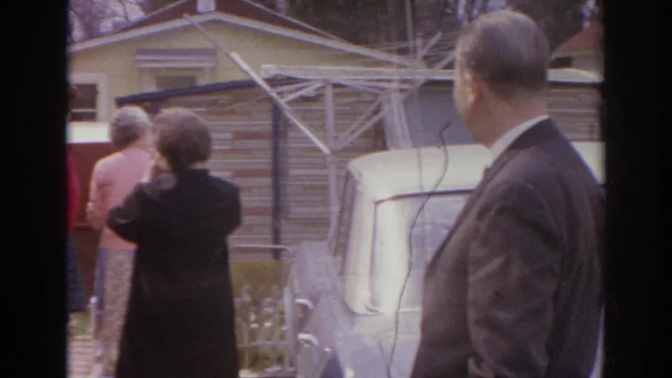 1966 Norfolk frame with a suited man in the foreground and adults conversing near a parked car in a residential yard.