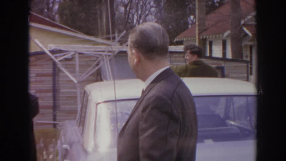 Home-movie frame from Norfolk in 1966 showing a suited man by a parked car with a residential house and yard structures behind him.