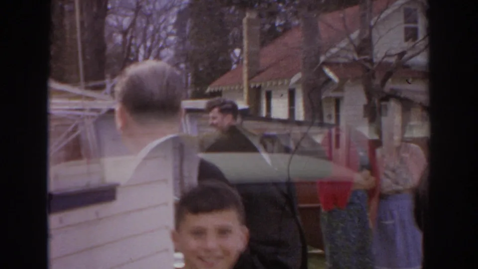 1966 Norfolk home-movie frame showing adults near a parked car and house, with a smiling boy visible in the foreground.