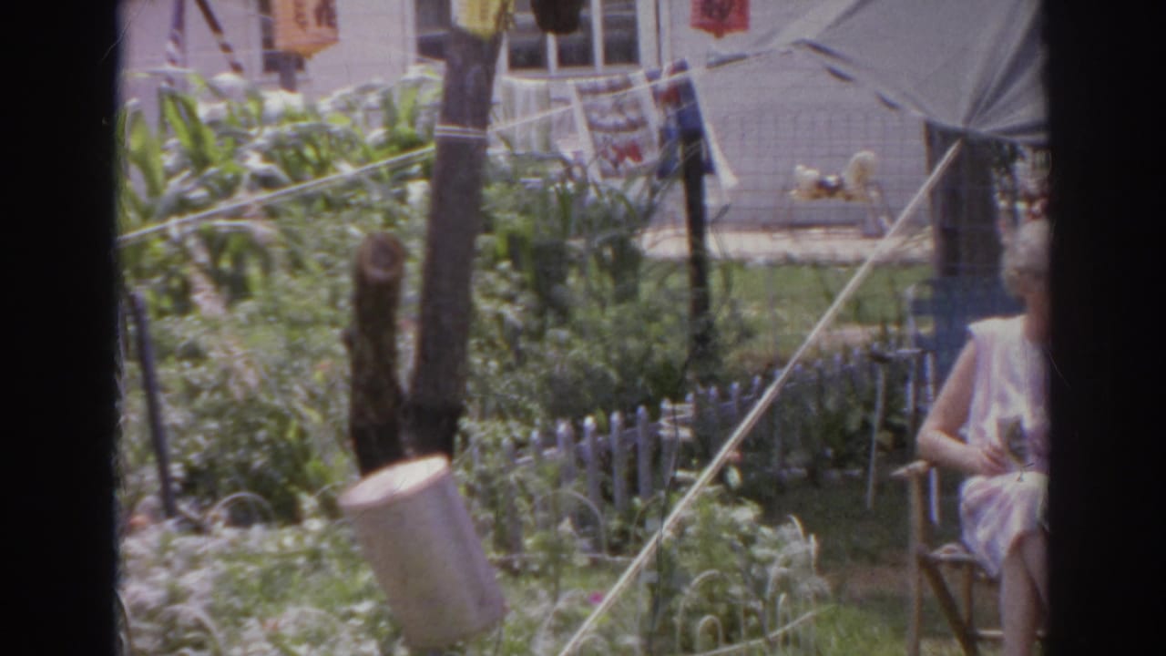 1966 Norfolk backyard frame showing a garden, lanterns, house wall, tent canopy, and a woman seated or standing partly outside the right edge.