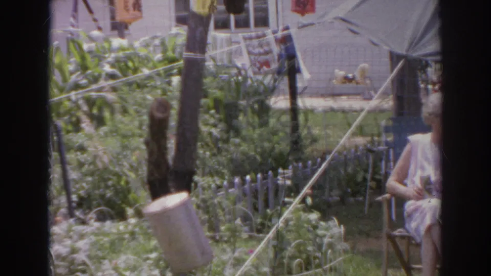 1966 Norfolk backyard frame showing a garden, lanterns, house wall, tent canopy, and a woman seated or standing partly outside the right edge.