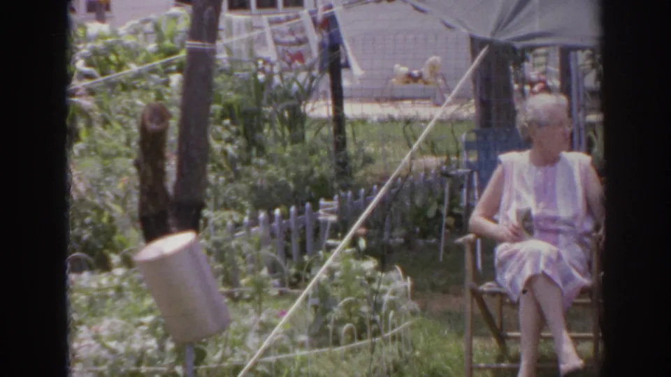 1966 Norfolk home-movie frame of an elderly woman in a light dress seated in a wooden chair in a backyard garden, viewed through window framing.