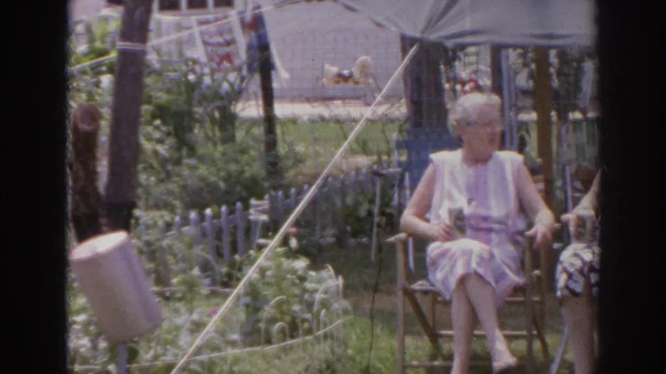1966 Norfolk backyard frame showing an elderly woman seated in a light dress while another woman is partly visible beside her in the garden setting.