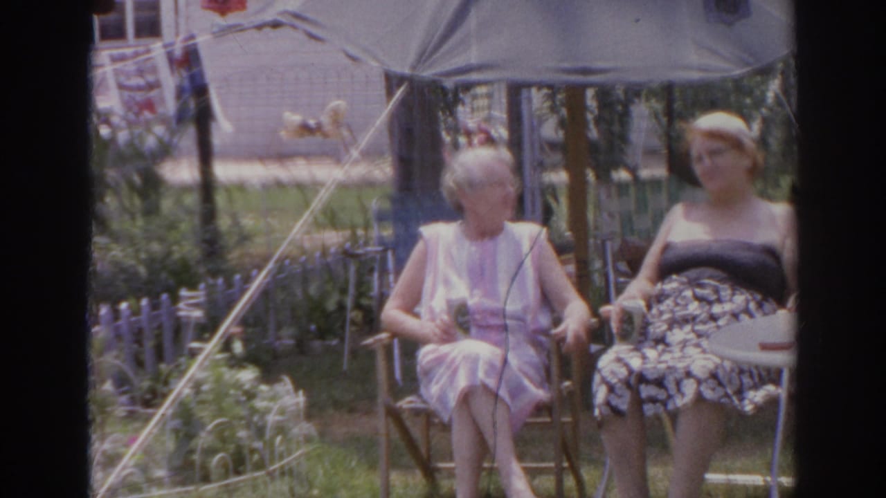 Two older women relax beneath a backyard umbrella in 1966 Norfolk, with laundry and fencing behind them.